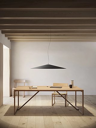 Eine breite, schwarze Pendelleuchte hängt über einem Holztisch in einem minimalistischen Raum.
