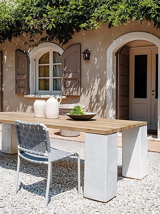 Ein Holztisch mit Stühlen auf einer Schotterterrasse vor einem Haus mit Fensterläden.