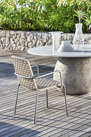 Ein geflochtener Stuhl vor einem runden Steintisch auf einer Holzterrasse.