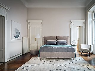 Ein Bett mit beigem Stoffbezug in einem Schlafzimmer im Altbau.