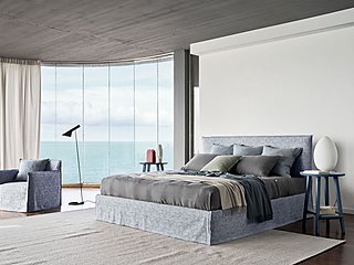 Ein blau-graues Bett vor einem großen Fenster mit Meerblick.