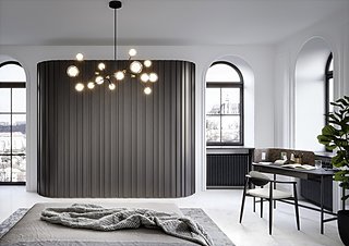 Ein Bett mit grauem Bezug vor einem dunkelgrauen, gerippten Kleiderschrank in einem Schlafzimmer mit Bogenfenstern.