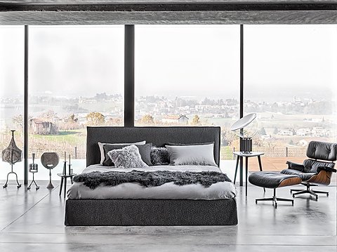 Ein dunkles Bett mit einem schwarzen Ledersessel in einem Raum mit Blick ins Grüne.