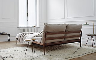 Ein Bett mit Holzrahmen und beigen Kissen am Kopfteil in einem weißen Raum.