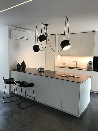 Weiße Küche mit Kochinsel, schwarzen Pendelleuchten und Barhockern auf Betonboden.