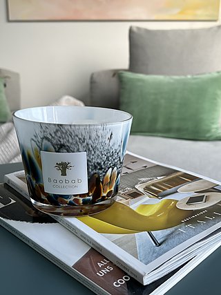 Duftkerze von Baobab auf Zeitschriften vor einem Sofa mit grünen Kissen.