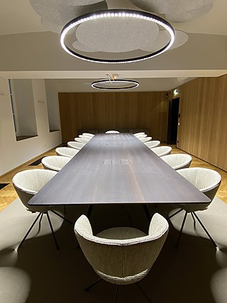 Konferenzraum mit langem dunklem Holztisch, hellen Polsterstühlen und runder Deckenleuchte.