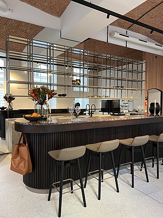 Geschwungenes Barmöbel aus dunklem Holz mit Steinplatte, Barhockern und Metallregal dahinter.