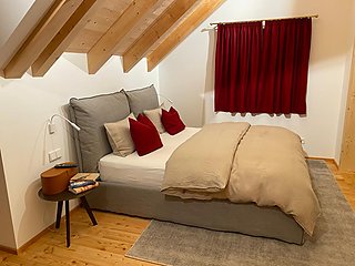 Schlafzimmer im Dachgeschoss mit Polsterbett, rotem Vorhang und Holzbalken.