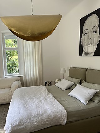 Schlafzimmer mit Polsterbett in Salbeigrün, goldener Pendelleuchte in Halbmondform und großem Porträt.