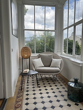 Geflochtener Sessel mit Kissen im Wintergarten mit Sprossenfenstern und Zementfliesen.