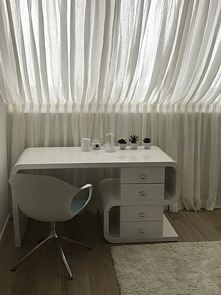 Weißer Schreibtisch mit Schubladen und Drehstuhl vor bodenlangen Vorhängen.